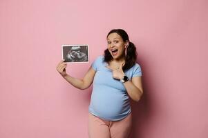 lycklig härlig gravid kvinna poäng åt sidan de ultraljud skanna bild av henne framtida bebis, isolerat på rosa bakgrund, foto