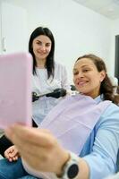 Lycklig gravid kvinna leende ser på henne spegel reflexion, Sammanträde i tandläkarens stol medan en dental utnämning foto