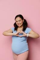 glad gravid kvinna gester med händer, som visar hjärta form med fingrar, ler ser på kamera, isolerat på rosa foto