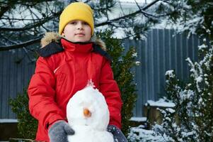 stilig förtjusande barn pojke 10 år gammal, i värma röd ner jacka, spelar med en snögubbe på en snö täckt bakgård foto