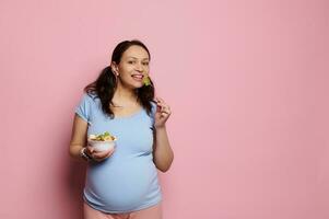 vackert gravid kvinna leende på kamera, innehav en skål med friska frukter, isolerat på rosa bakgrund. kopia Plats foto