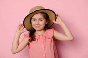 äkta liten barn flicka i rosa klänning, ler ser på kamera, sätta på en eleganta sugrör hatt, isolerat bakgrund foto