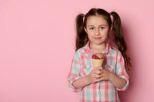 skön liten unge flicka med två hästsvansar, innehav en våffla is grädde, leende ser på kamera, isolerat på rosa foto