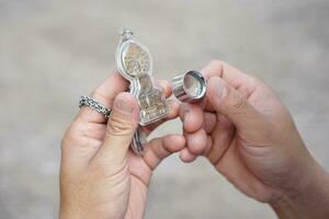 närbild händer håll buddha amulett och förstorande glas till kolla upp detaljer och figur av objekt. begrepp, tro och tro för buddist för helig och tur. insamling amulett som hobbyer, utbyta, försäljning. foto