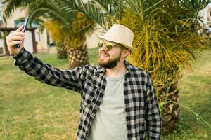 man tar selfie porträtt över handflatan träd bakgrund - Lycklig tusenåriga kille njuter sommar högtider i stad - ungdom och teknik foto