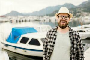 stilig man bär hatt och glasögon nära marina med yachter. porträtt av skrattande man med hav hamn bakgrund med kopia Plats foto