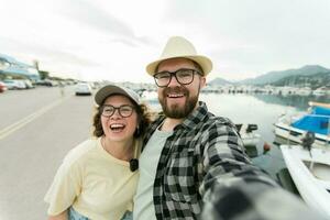 ung par tar en själv porträtt skrattande som de utgör stänga tillsammans för kamera på deras smartphone utomhus i sommar hamn marina med båtar och yachter foto