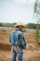 cowboy bär jeans och en hatt, stående under de himmel med fluffig moln, arbetssätt på en bruka foto