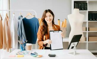 asiatisk skräddare kvinna arbetssätt på kläder i skrädderi ateljé. skön ung kvinna mode designer leende och efter Framgång i studio foto