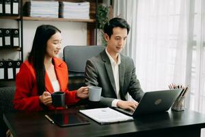 två asiatisk företag arbetare talande på de smartphone och använder sig av bärbar dator medan Sammanträde i kontor rum. foto