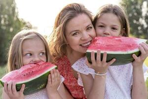 porträtt av mamma med söt döttrar ha roligt medan äter en skiva av vattenmelon foto