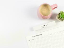 platt lägga av vana tracker bok, trä- kalender juli, rosa kopp av svart kaffe och saftig växt pott på vit bakgrund med kopia Plats. foto
