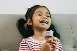Lycklig asiatisk liten flicka äter is grädde på soffa på Hem. foto