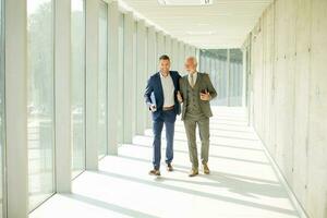 ung och en senior affärsman promenad ner ett kontor hall, djup i konversation foto
