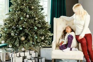 Lycklig familj kvinna mor och liten flicka koppla av spelar sparkler nära jul träd på jul eve på Hem. mamma, dotter i hus med vinter- dekoration. jul ny år tid för firande foto