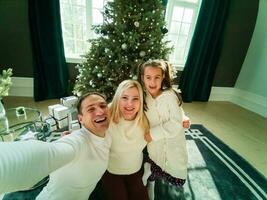 familj, högtider, teknologi och människor - leende mor, far och liten flicka framställning selfie med kamera över levande rum och jul träd bakgrund foto