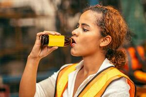afrikansk amerikan kvinna arbetstagare dricka energi dryck under koppla av broms arbete i tung fabrik. foto
