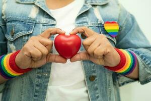 asiatisk dam som bär regnbågsflaggarmband och håller rött hjärta, symbol för HBT stolthet månad firar årliga i juni sociala av homosexuella, lesbiska, bisexuella, transpersoner, mänskliga rättigheter. foto