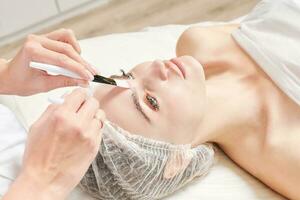 kosmetolog gnuggning grädde med borstar in i kvinna ansikte hud för föryngring, kosmetisk procedur foto