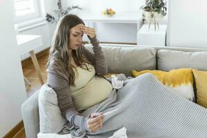 ung gravid kvinna liggande i säng, mätning kropp temperatur, innehav termometer och ansikte mask foto