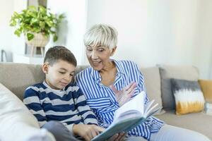 kärleksfull mormor undervisning barnbarn innehav bok Sammanträde på soffa, mormor bebis barnvakt fattande unge pojke läsning saga till söt barn, barnskötare mormor talande berättelse till förskola barnbarn foto