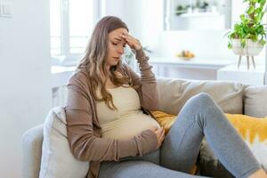 gravid ung vuxen kvinna vilar på soffa på Hem, känsla sjuk. ung gravid kvinna har lidit från huvudvärk Sammanträde på de soffa. graviditet symptom foto