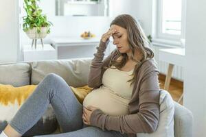 gravid ung vuxen kvinna vilar på soffa på Hem, känsla sjuk. ung gravid kvinna har lidit från huvudvärk Sammanträde på de soffa. graviditet symptom foto