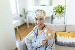 senior kvinna ser Lycklig efter få vaccin. mogna kvinna med plåster på henne ärm efter tar emot covid-19 vaccination Sammanträde på Hem foto