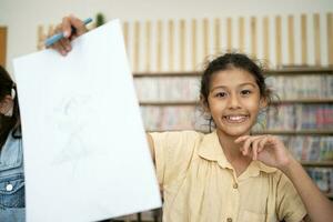 porträtt av leende liten elev skrivning på skrivbord i klassrum på de elementärt skola. studerande flicka håller på med testa i primär skola. barn skrivning anteckningar i klassrum. utbildning kunskap begrepp foto