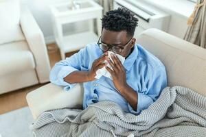 sjuk afrikansk ung man täckt med filt blåser löpning näsa fick feber fångad kall nysning i vävnad sitta på soffa, sjuk allergisk svart man har allergi symptom hosta på Hem, korona 19 foto