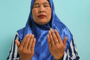 närbild se av en medelålders asiatisk kvinna i en blå hijab och randig skjorta är framställning en bön- gest med henne händer Uppfostrad upp. hon är isolerat över en blå bakgrund foto