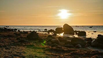 solnedgång, sten strand med små och stor stenar i främre av de upplyst hav. foto