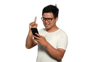 upphetsad asiatisk man bär vit tshirt leende medan innehav hans telefon, isolerat förbi vit bakgrund foto