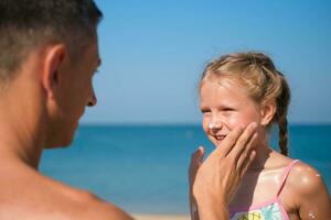 far applicering skyddande grädde till henne dotters ansikte på de strand. man hand innehav Solskydd lotion på bebis ansikte. söt liten flicka med en Sol blockera förbi de hav . kopia Plats. foto