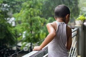 liten unge spelar i sommar regn i hus balkong, indisk smart pojke spelar med regn droppar under monsun regnig säsong, unge spelar i regn foto