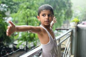liten unge spelar i sommar regn i hus balkong, indisk smart pojke spelar med regn droppar under monsun regnig säsong, unge spelar i regn foto