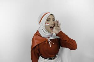 ung skön muslim kvinna bär en röd topp och vit hijab är innehav Indonesiens flagga och skrikande och skrikande högt med en hand på henne mun. Indonesiens oberoende dag begrepp. foto