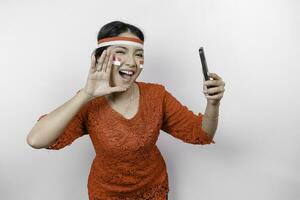 ung skön kvinna bär en röd kebaya är innehav henne telefon medan skrikande och skrikande högt med en hand på henne mun. Indonesiens oberoende dag begrepp. foto