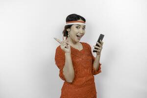 en Lycklig asiatisk kvinna bär röd kebaya och pannband, innehav henne telefon, och pekande kopia Plats på topp av henne, isolerat förbi vit bakgrund. Indonesiens oberoende dag foto