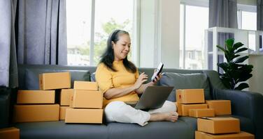 mogna företag ägare kvinna förbereda paket låda och stående kolla upp uppkopplad order för leverera till kund på läsplatta, bärbar dator handla uppkopplad foto
