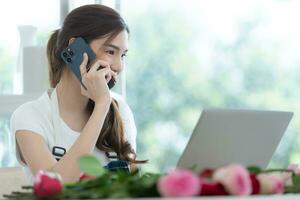 skön asiatisk kvinna talande med kund på de telefon och använder sig av bärbar dator på blommig affär. foto