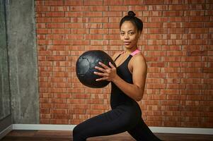 sida porträtt av ett attraktiv muskulös bygga afrikansk amerikan kvinna i eleganta svart sportkläder, utövar med medicin boll på Gym, sött ler ser på kamera foto