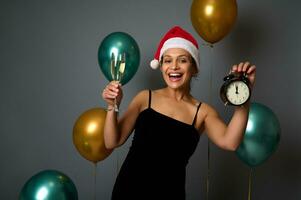 skön kvinna i santa karneval hatt ler toothy leende Framställ mot skinande luft ballonger på grå bakgrund med larm klocka och champagne glas i händer, kopia ad Plats. jul, Lycklig ny år foto