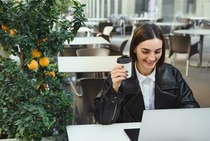 skön ung företag kvinna, frilansare, utvecklaren, journalist, copywriter innehar hämtmat kopp av kaffe, Arbetar uppkopplad i utomhus- Kafé, ler läsning Bra Nyheter från företag partners på bärbar dator foto