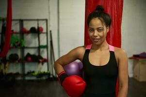 ung kvinna boxare, sportig flicka, idrottare kvinna, sporter kvinna, bär röd boxning handskar, utseende på kamera Framställ mot sporter Utrustning på de Gym foto
