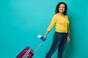 Lycklig lockigt mörkhårig kvinna, resande i ljus gul Tröja och blå jeans innehav pass med ombordstigning passera och luft biljett, ler, Framställ med bagage på blå bakgrund med kopia Plats foto