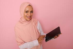 arab muslim kvinna i rosa hijab ler med toothy leende ser på kamera, Framställ på färgad pastell bakgrund med digital läsplatta i händer. isolerat porträtt för reklam med kopia Plats för text foto