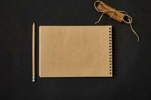 platt lägga minimalistisk sammansättning av en hantverk anteckningsbok med tömma tom papper ark med kopia Plats, en trä- penna och bunden kanel med en rep på svart bakgrund foto