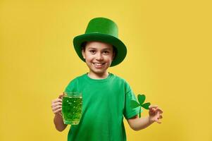 liten pojke bär pyssling irländsk hatt innehar en glas med grön dryck och en klöver blad ler medan Framställ på kamera. helgon Patricks dag foto