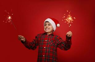 jul och ny år firande begrepp. rolig stilig barn pojke i santa hatt med sparklers mot röd färgad bakgrund med kopia Plats för ad foto